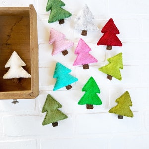 May include: Ten felt Christmas tree ornaments in various colors, including green, red, pink, white, and blue. The ornaments are arranged on a white brick background.