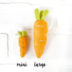 May include: Two felt carrots, one mini and one large, on a white brick background. The carrots are orange with green tops.