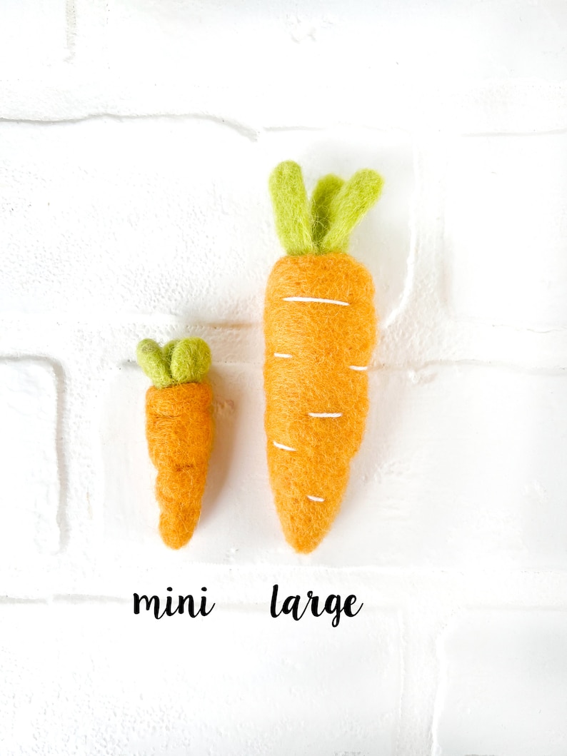 May include: Two felt carrots, one mini and one large, on a white brick background. The carrots are orange with green tops.