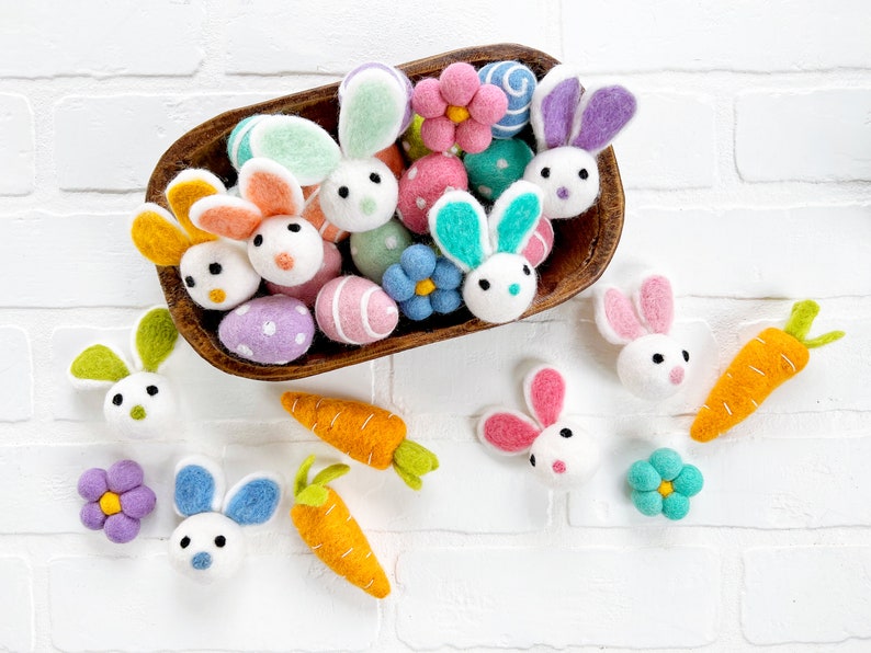 May include: A wooden bowl filled with felt Easter decorations, including bunnies, eggs, flowers, and carrots. The decorations are in various colors, including pink, blue, green, yellow, and purple.