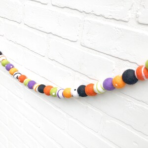 May include: A colorful Halloween garland featuring felt balls in various colors, including black, orange, purple, green, and white. The garland is strung together, creating a festive decoration for the holiday season.