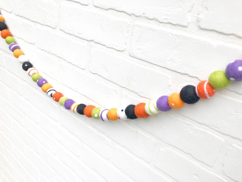 May include: A colorful Halloween garland featuring felt balls in various colors, including black, orange, purple, green, and white. The garland is strung together, creating a festive decoration for the holiday season.