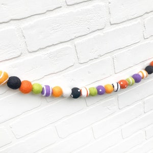 May include: A colorful Halloween felt ball garland. The garland features a variety of felt balls in orange, black, white, purple, green, and white with orange stripes. The garland is strung together and is displayed against a white brick wall.