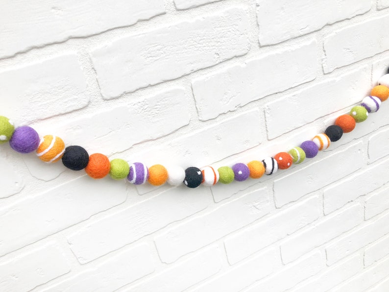 May include: A colorful Halloween felt ball garland. The garland features a variety of felt balls in orange, black, white, purple, green, and white with orange stripes. The garland is strung together and is displayed against a white brick wall.