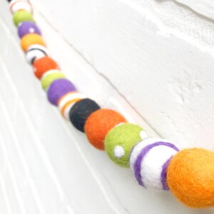 May include: A colorful felt ball garland with orange, black, white, green, and purple balls. The balls are strung together, creating a festive decoration. The garland is displayed against a white brick wall.