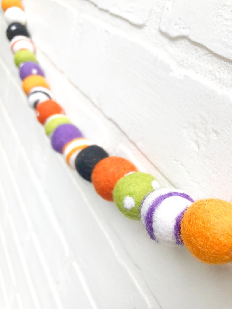 May include: A colorful felt ball garland with orange, black, white, green, and purple balls. The balls are strung together, creating a festive decoration. The garland is displayed against a white brick wall.