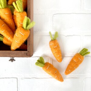 May include: A wooden box overflowing with felt carrots, with three additional carrots spilling out onto a white brick background. The carrots are orange with green tops and white stitching.