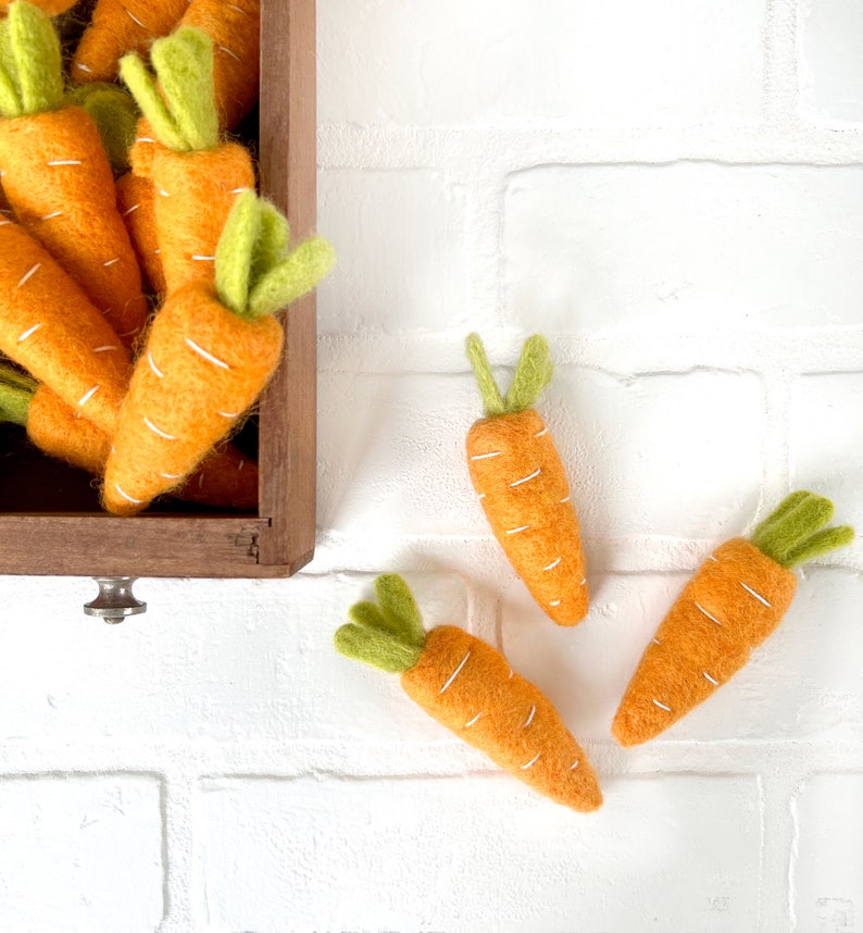 May include: A wooden box overflowing with felt carrots, with three additional carrots spilling out onto a white brick background. The carrots are orange with green tops and white stitching.