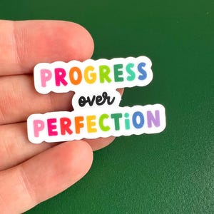 Puede incluir: Una pegatina arcoíris con el texto "PROGRESS over PERFECTION" en una fuente divertida.