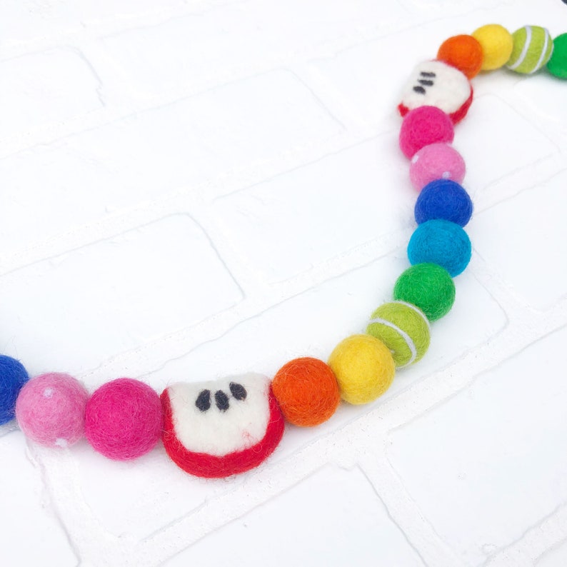 Puede incluir: Una colorida guirnalda de bolas de fieltro con un arco&iacute;ris de tonos, incluyendo rojo, naranja, amarillo, verde, azul y rosa. La guirnalda presenta bolas de fieltro y rodajas de manzana, ideal para decoraci&oacute;n.