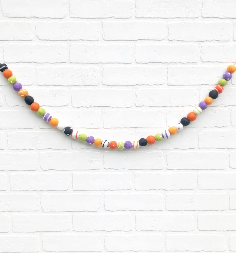 May include: A colorful Halloween garland featuring felt balls in orange, black, purple, green, and white. The balls are decorated with stripes, polka dots, and other patterns. The garland is strung on a white string and is displayed against a white brick wall.