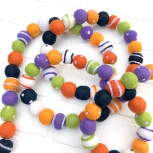 May include: A colorful felt ball garland in a variety of colors, including orange, purple, green, black, and white. Some balls have stripes or polka dots. The garland is likely for festive or decorative purposes.