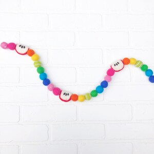 Puede incluir: Una colorida guirnalda de bolas de fieltro con un arco&iacute;ris de colores, incluyendo rosa, naranja, amarillo, verde y azul. La guirnalda tambi&eacute;n presenta rodajas de manzana de fieltro con detalles blancos y negros. Es un art&iacute;culo decorativo.