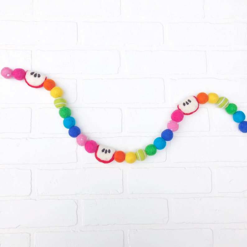 Puede incluir: Una colorida guirnalda de bolas de fieltro con un arco&iacute;ris de colores, incluyendo rosa, naranja, amarillo, verde y azul. La guirnalda tambi&eacute;n presenta rodajas de manzana de fieltro con detalles blancos y negros. Es un art&iacute;culo decorativo.