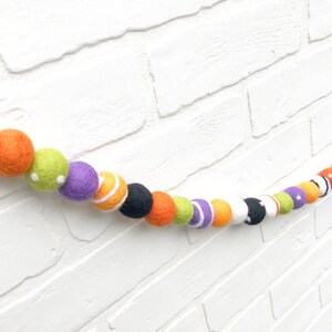 May include: A felt ball garland with a Halloween theme. The garland features a variety of colorful felt balls in orange, black, white, green, and purple. Some balls have stripes, dots, or other patterns. The garland is displayed against a white brick wall.