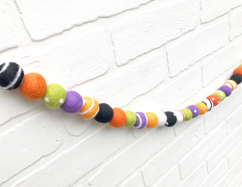 May include: A felt ball garland with a Halloween theme. The garland features a variety of colorful felt balls in orange, black, white, green, and purple. Some balls have stripes, dots, or other patterns. The garland is displayed against a white brick wall.