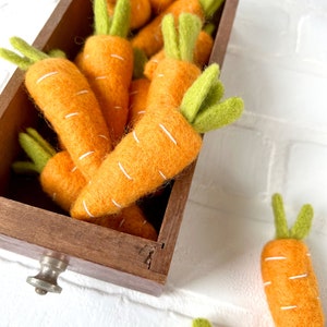 May include: A wooden box filled with felt carrots. The carrots are orange with white stitching and green felt tops. Two additional carrots are on a white surface next to the box.