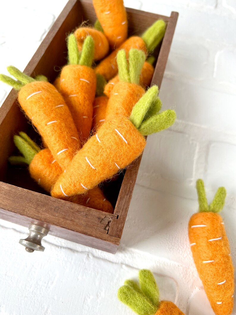 May include: A wooden box filled with felt carrots. The carrots are orange with white stitching and green felt tops. Two additional carrots are on a white surface next to the box.