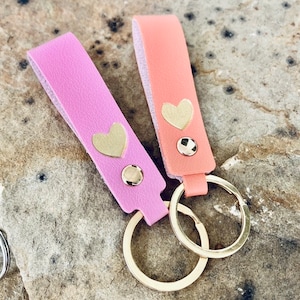 May include: Two leather keychains with gold heart charms and gold key rings. One keychain is pink and the other is peach.