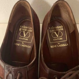 May include: A pair of brown leather dress shoes with laces. The shoes have a suede upper and a leather toe cap. The insole features the "Valentino Garavani Couture by Rene Caovilla" logo in gold lettering.