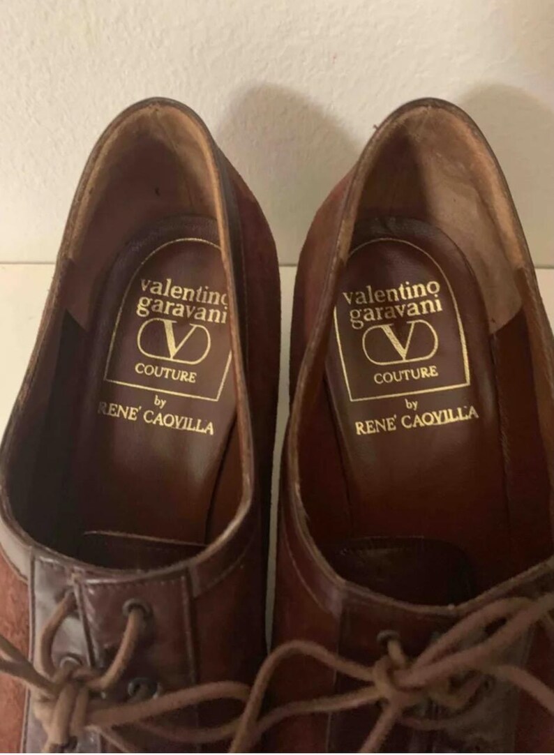 May include: A pair of brown leather dress shoes with laces. The shoes have a suede upper and a leather toe cap. The insole features the "Valentino Garavani Couture by Rene Caovilla" logo in gold lettering.