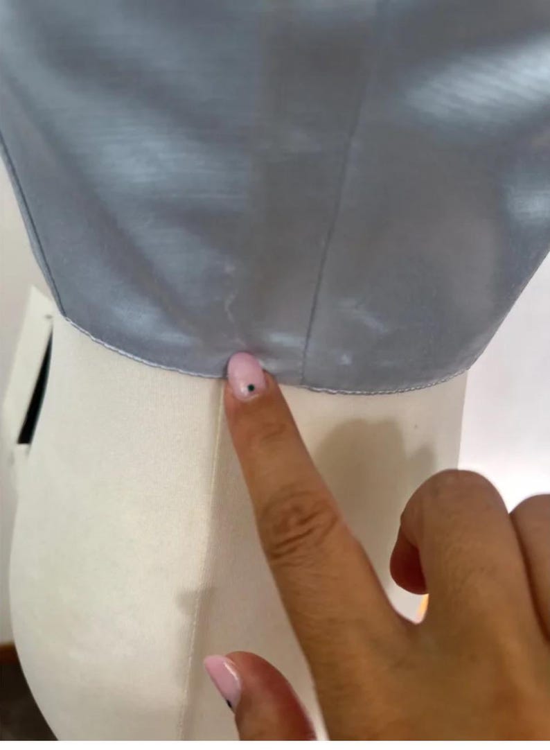 Puede incluir: Primer plano del borde inferior de una tela de sat&eacute;n azul claro. La tela es lisa y tiene un ligero brillo. La tela est&aacute; siendo sostenida por una mano.