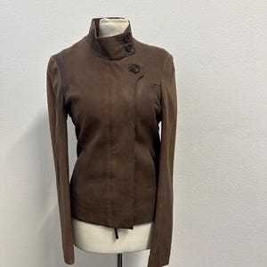 May include: A brown leather jacket with a high collar and long sleeves. The jacket has a button closure and is displayed on a mannequin. The jacket's color is a rich, earthy brown, and the design is minimalist and modern.