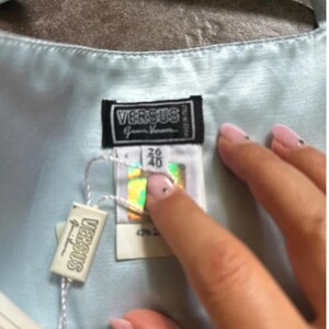 Puede incluir: Una blusa de sat&eacute;n azul claro con una etiqueta blanca que dice "Versus" y una etiqueta blanca con el texto "Versus Gianni Versace" y la talla 36/40.