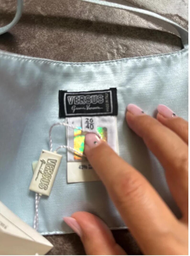 Puede incluir: Una blusa de sat&eacute;n azul claro con una etiqueta blanca que dice "Versus" y una etiqueta blanca con el texto "Versus Gianni Versace" y la talla 36/40.