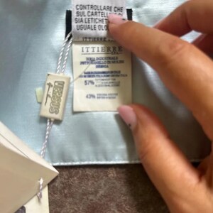 Puede incluir: Una bufanda de seda azul claro con una etiqueta blanca adjunta. La etiqueta tiene el texto "ITTIERRE" y "CONTROLLARE CHE SUL CARTELL SIA LETICHET UGUALE OLO" impreso en ella. La etiqueta tambi&eacute;n tiene el texto "VERSUS" impreso en ella.