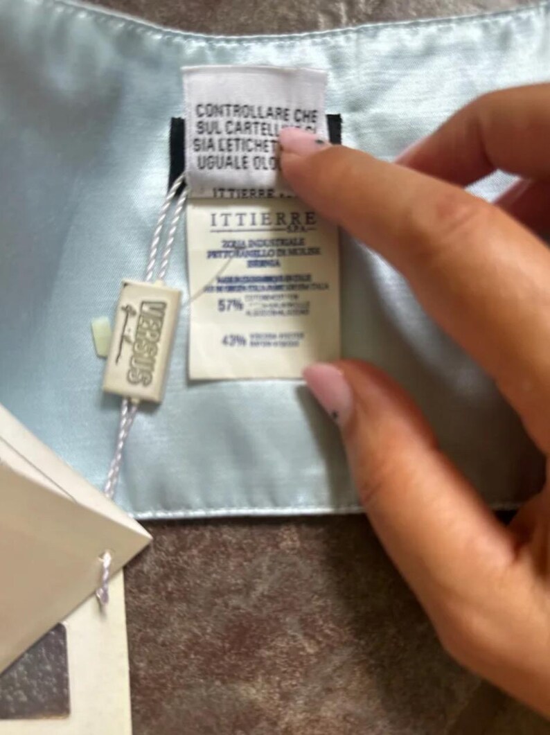Puede incluir: Una bufanda de seda azul claro con una etiqueta blanca adjunta. La etiqueta tiene el texto "ITTIERRE" y "CONTROLLARE CHE SUL CARTELL SIA LETICHET UGUALE OLO" impreso en ella. La etiqueta tambi&eacute;n tiene el texto "VERSUS" impreso en ella.