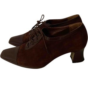 May include: A pair of brown suede and leather oxford-style shoes. The shoes have a pointed toe, a lace-up closure, and a small heel. The toe and heel are a darker shade of brown than the suede upper.