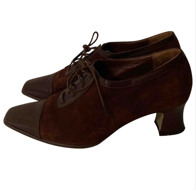 May include: A pair of brown suede and leather oxford-style shoes. The shoes have a pointed toe, a lace-up closure, and a small heel. The toe and heel are a darker shade of brown than the suede upper.