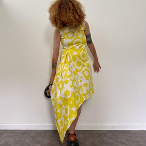 Può includere: Abito bianco senza maniche con motivo floreale giallo. L'abito ha un orlo asimmetrico e un corpetto aderente. La modella indossa scarpe nere con cinturini marroni. L'abito è in stile vintage.