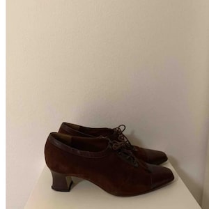 May include: A pair of brown suede and leather heeled shoes. The shoes have a lace-up design with a rounded toe and a small, dark heel. The shoes are displayed on a white surface.