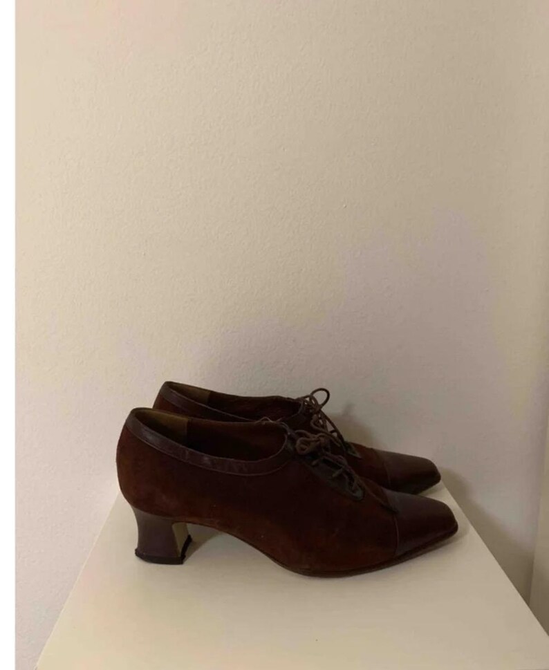 May include: A pair of brown suede and leather heeled shoes. The shoes have a lace-up design with a rounded toe and a small, dark heel. The shoes are displayed on a white surface.