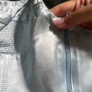 Puede incluir: Una blusa de sat&eacute;n azul claro con cierre de cremallera y un detalle de puntada decorativa.