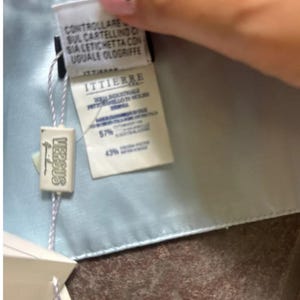 Puede incluir: Una tela de sat&eacute;n azul claro con una etiqueta blanca adjunta. La etiqueta tiene el texto "VERSACE" en letras negras y una etiqueta blanca con el texto "ITTIERE" en letras negras. La etiqueta blanca tiene el texto "CONTROLLARE SUL CARTELLINO SIA LETICHETTA CON UGUALE OLOGIRIFFE" en letras negras.