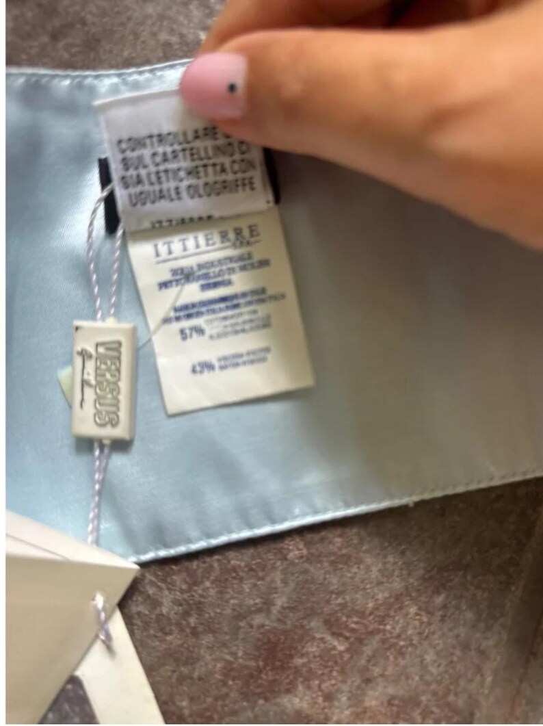 Puede incluir: Una tela de sat&eacute;n azul claro con una etiqueta blanca adjunta. La etiqueta tiene el texto "VERSACE" en letras negras y una etiqueta blanca con el texto "ITTIERE" en letras negras. La etiqueta blanca tiene el texto "CONTROLLARE SUL CARTELLINO SIA LETICHETTA CON UGUALE OLOGIRIFFE" en letras negras.