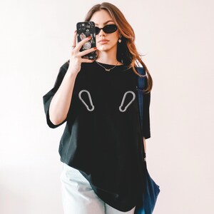 Peut inclure: Un t-shirt noir avec un motif de deux mousquetons argentés. La personne portant le t-shirt tient une coque de téléphone noire avec un motif coloré.