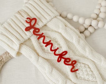 May include: Red acrylic stocking tag with the name "Jennifer" written in a cursive font. The tag is hanging on a white knitted stocking with a brown tassel.