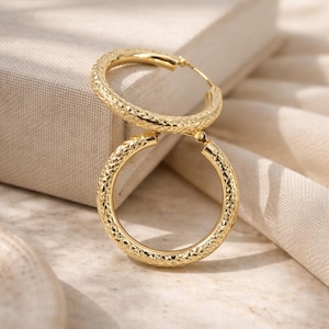 May include: A pair of gold hoop earrings featuring a textured, diamond-cut surface pattern. The circular earrings have a polished finish and a hinged closure mechanism, showcasing a reflective metallic sheen across the entire exterior surface.