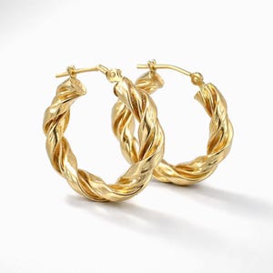 Puede incluir: Un par de pendientes de aro dorados con una textura trenzada similar a una cuerda. Los pendientes circulares tienen un acabado pulido y reflectante, e incluyen un mecanismo de poste con bisagra en la parte superior para un cierre seguro.