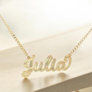 Puede incluir: Un collar dorado con el nombre "Julia" en fuente cursiva. El colgante está conectado a una cadena. El collar se muestra sobre una superficie de madera de color claro.