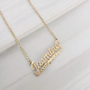 May include: A gold-colored necklace with the name "Jasmine" in a cursive font. The pendant has a textured, diamond-cut design. The chain is a curb link style, and the necklace is displayed on a white surface.
