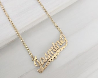 Può includere: Una collana color oro con il nome "Jasmine" in corsivo. Il ciondolo ha un design strutturato con taglio a diamante. La catena è in stile a maglia curb, esposta su una superficie bianca.