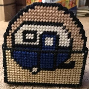 May include: A beige and blue crocheted coaster with a design of a robot face. The robot has black eyes, a white mouth, and a blue body.