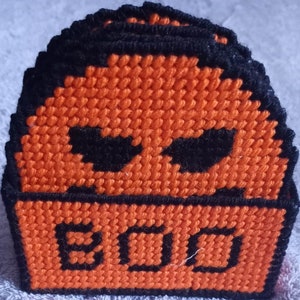 Puede incluir: Un juego de seis posavasos de crochet naranja y negro con un diseño de calavera y la palabra "BOO" en la parte inferior.