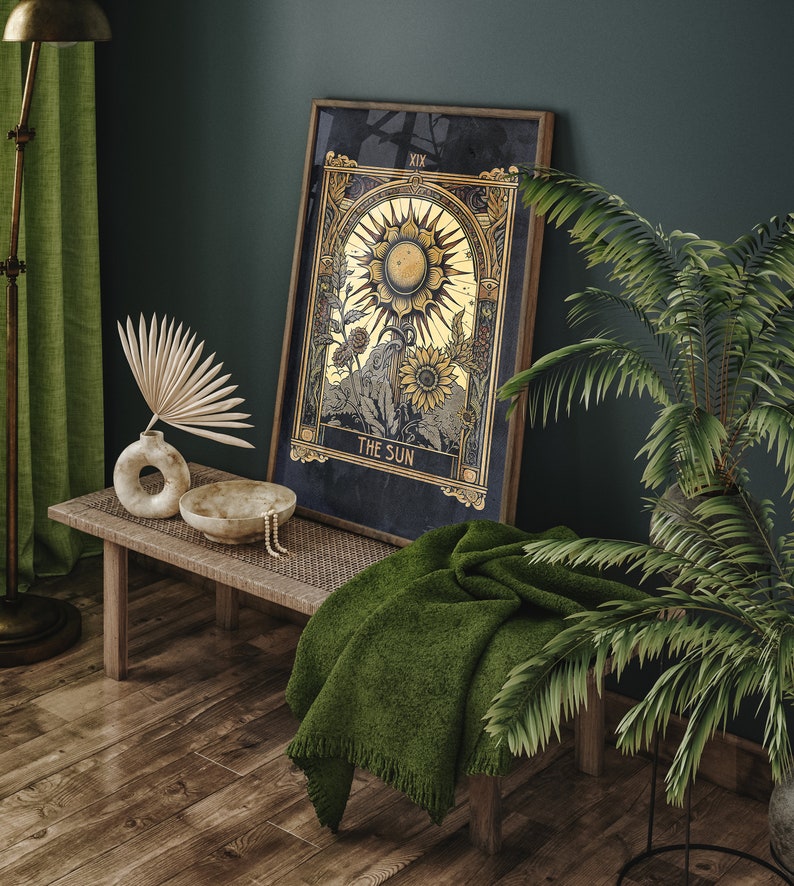 May include: A framed tarot card print with a gold and black design depicting the Sun card. The card is hanging on a dark green wall above a wooden bench with a green throw blanket. A potted plant with green leaves is in the corner of the image.