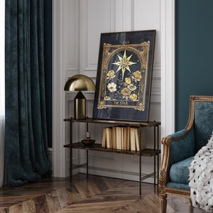 May include: A gold framed tarot card print with a dark blue background. The card depicts a star with yellow flowers and the text "XVII THE STAR". The print is hanging on a wall above a shelf with books and a lamp.